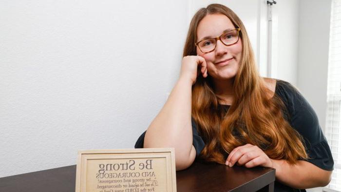 A student sits at a table, smiling with their head propped on their hand.